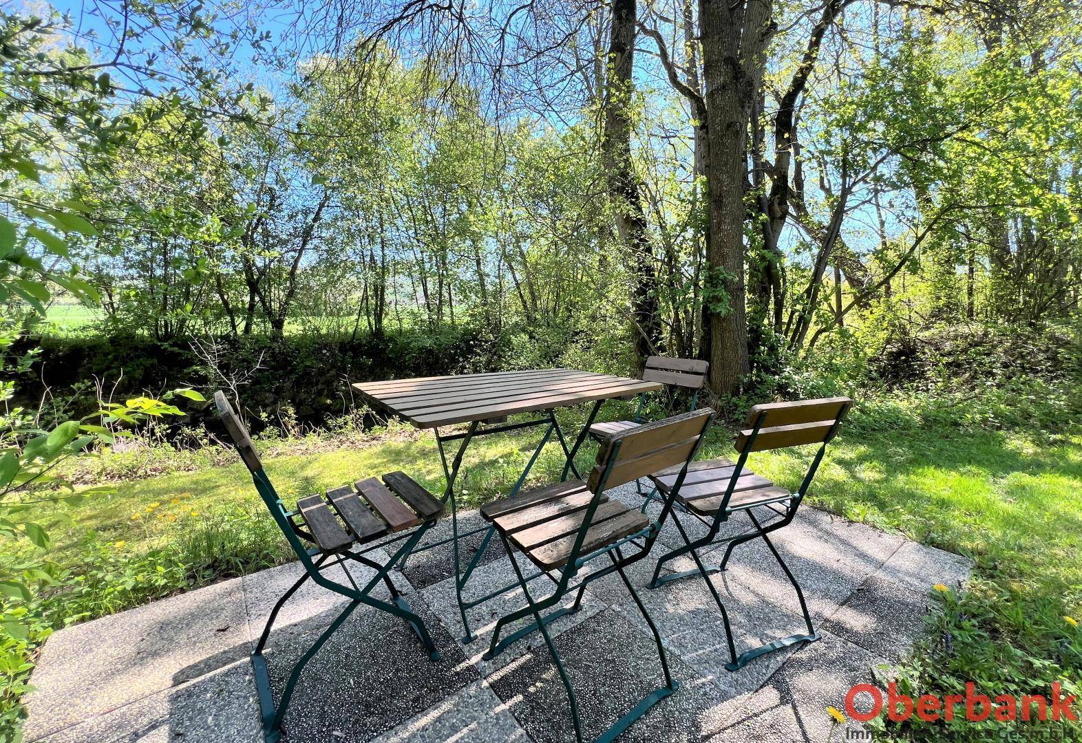 Gemütliche Terrasse mit Holztisch und Stühlen, umgeben von üppigem Grün und Bäumen, ideal zum Entspannen.
