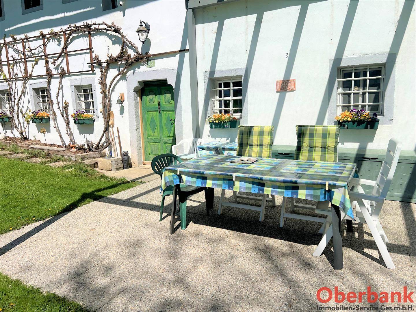 Sonniger Terrassenbereich mit Gartentisch und Stühlen, umgeben von grüner Natur und dem historischen Gebäude.