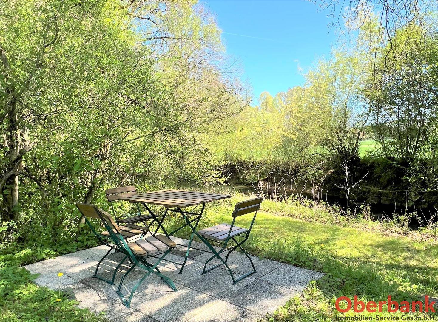 Idyllische Terrasse mit Gartenmöbeln, umgeben von Natur und einem kleinen Bach im Hintergrund, unter blauem Himmel.