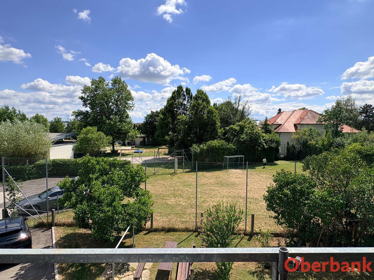 Weitläufiger Blick auf eine grüne Wiese mit Spielplatz und Bäumen unter blauem Himmel.