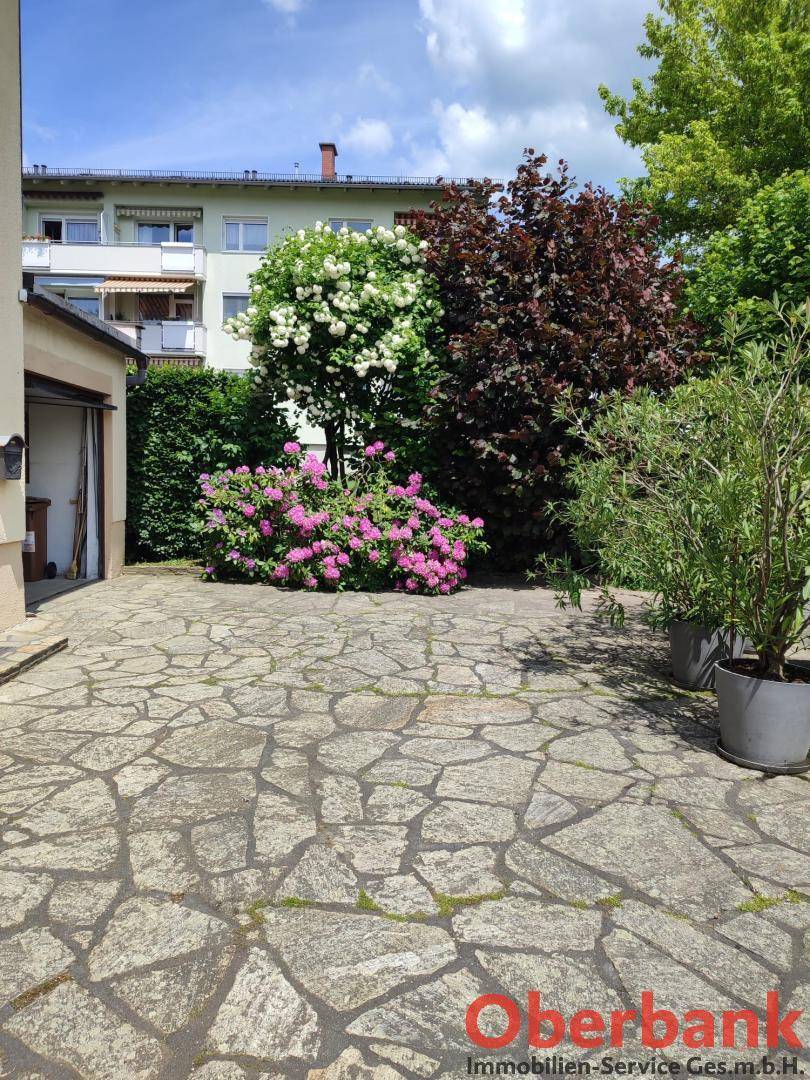 Gemütlicher gepflasterter Terrassenbereich mit Topfpflanzen und blühenden Sträuchern im Hintergrund.