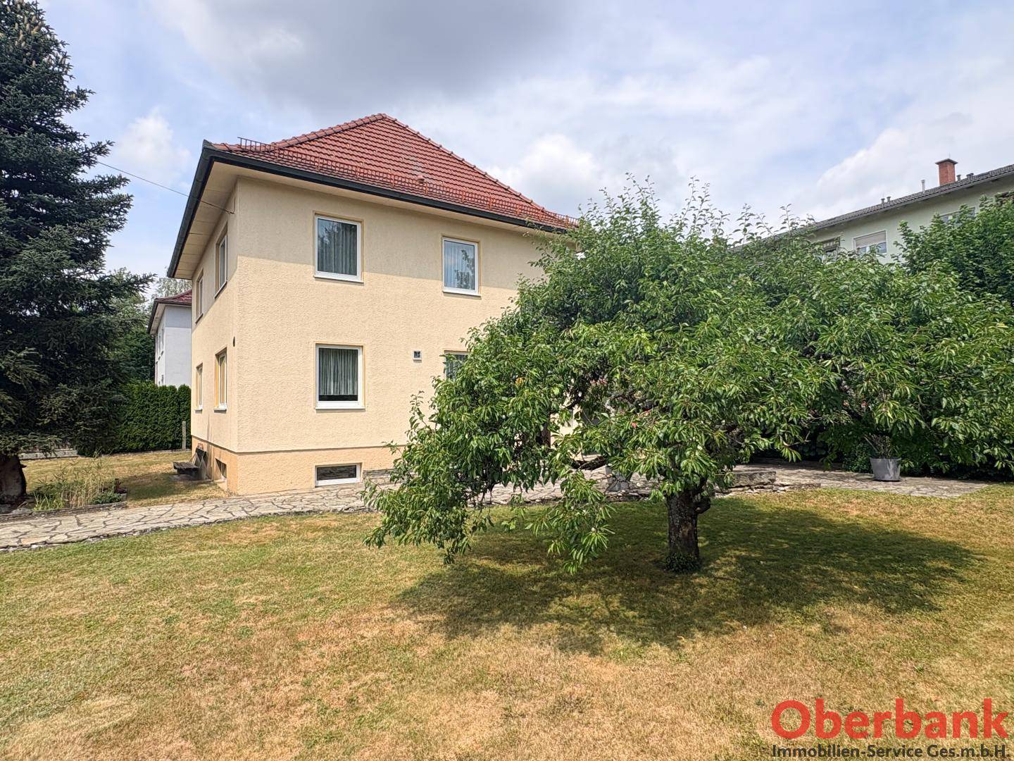 Ansicht des Hauses von der Seite mit einem großen, schattenspendenden Baum und weitläufigem Rasen.