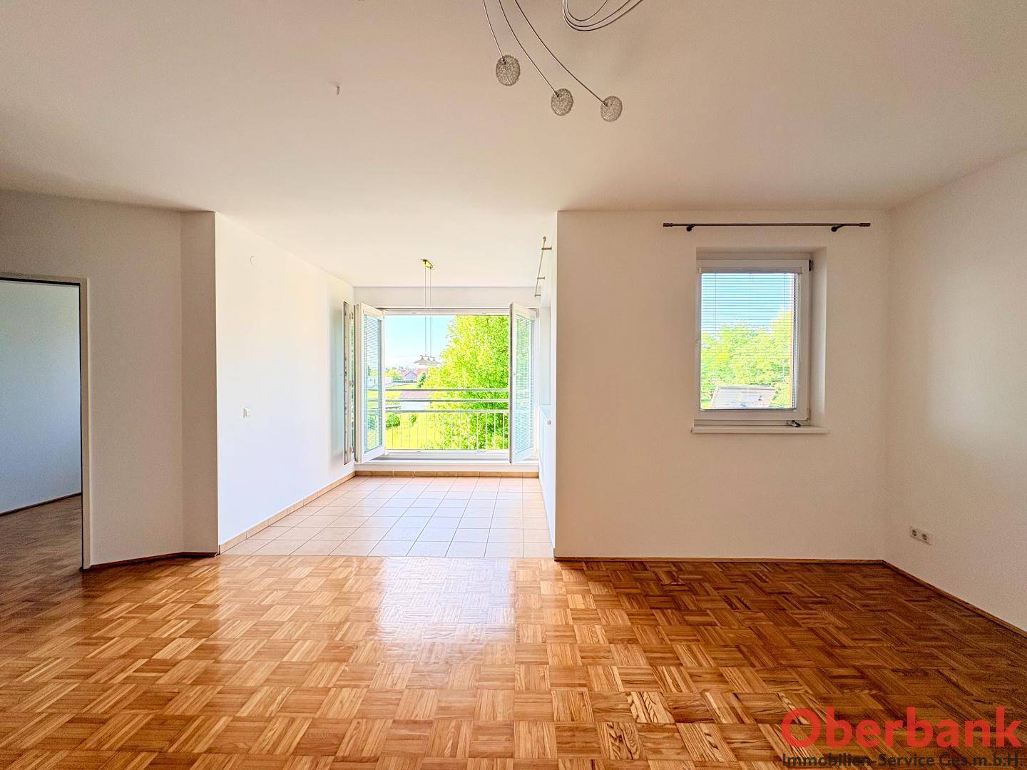 Geräumiges Wohnzimmer mit Parkettboden und Zugang zu einem hellen, gefliesten Bereich mit Balkonblick.