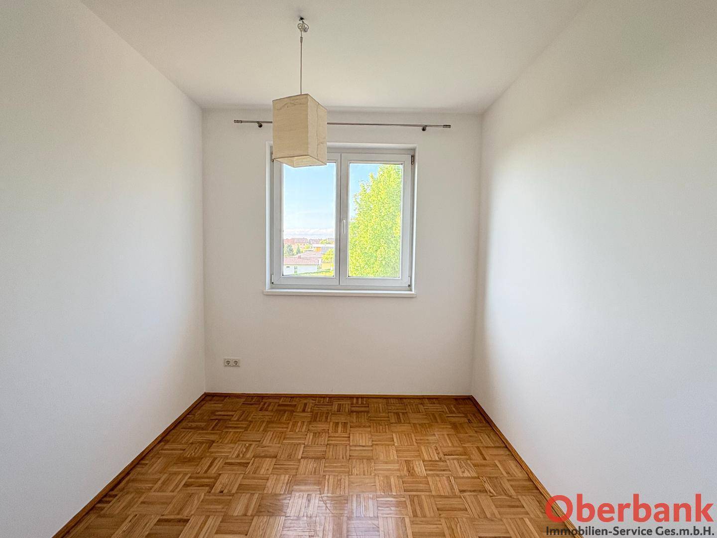 Helles Schlafzimmer mit Parkettboden und einem Fenster, das den Blick ins Grüne freigibt.