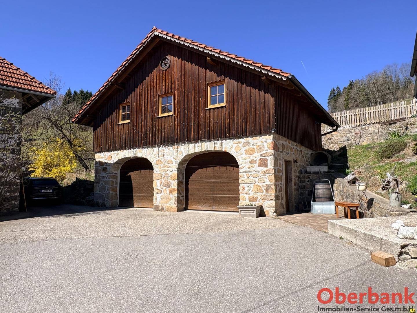 Traditionelles Gebäude mit zwei Garagentoren und Holzbalkon in ländlicher Idylle, umgeben von Grün.
