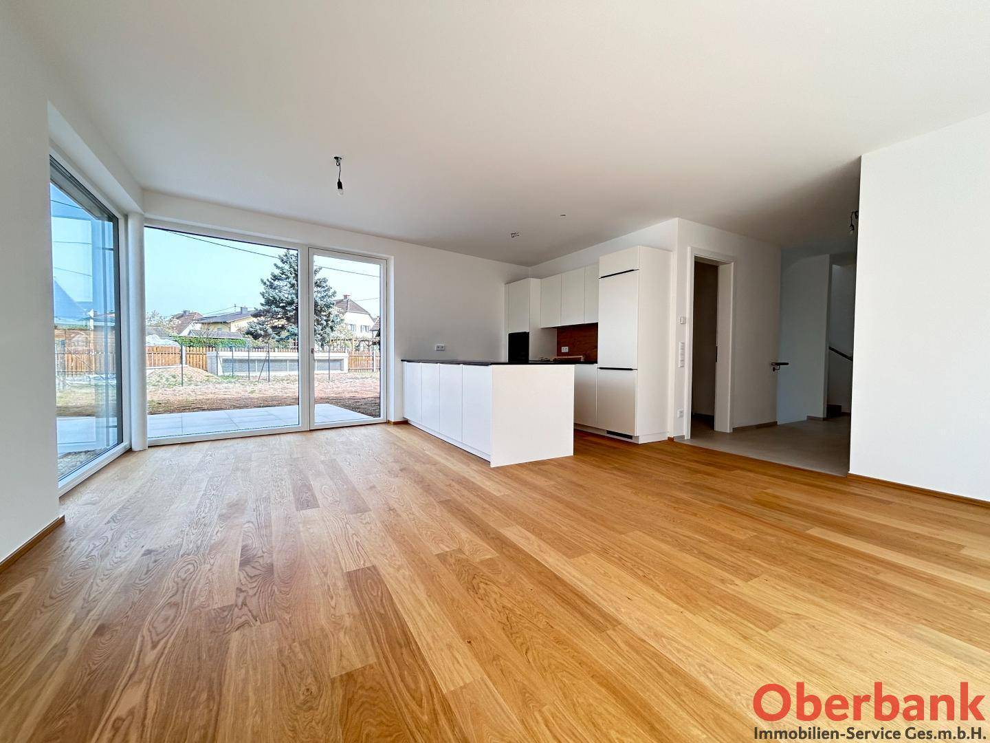 Offener Wohnbereich mit Holzboden, moderner weißer Küche und großen Fenstern zum Garten.