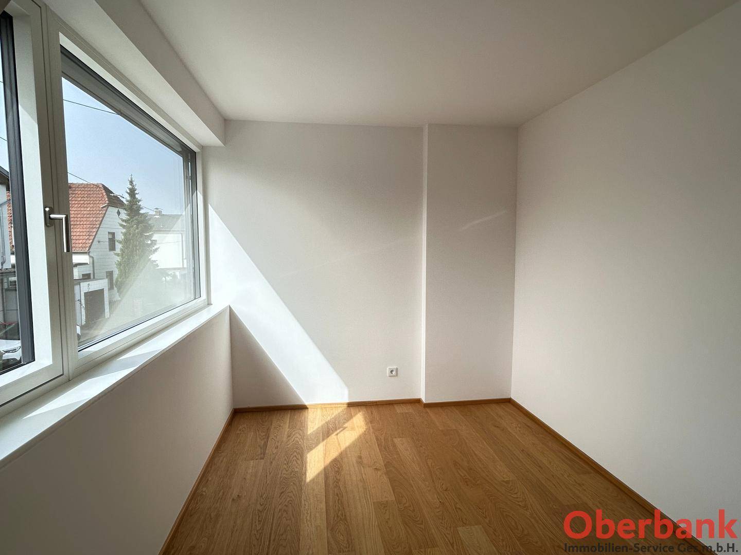 Heller Raum mit Holzboden und großem Fenster, das viel Sonnenlicht hereinlässt.