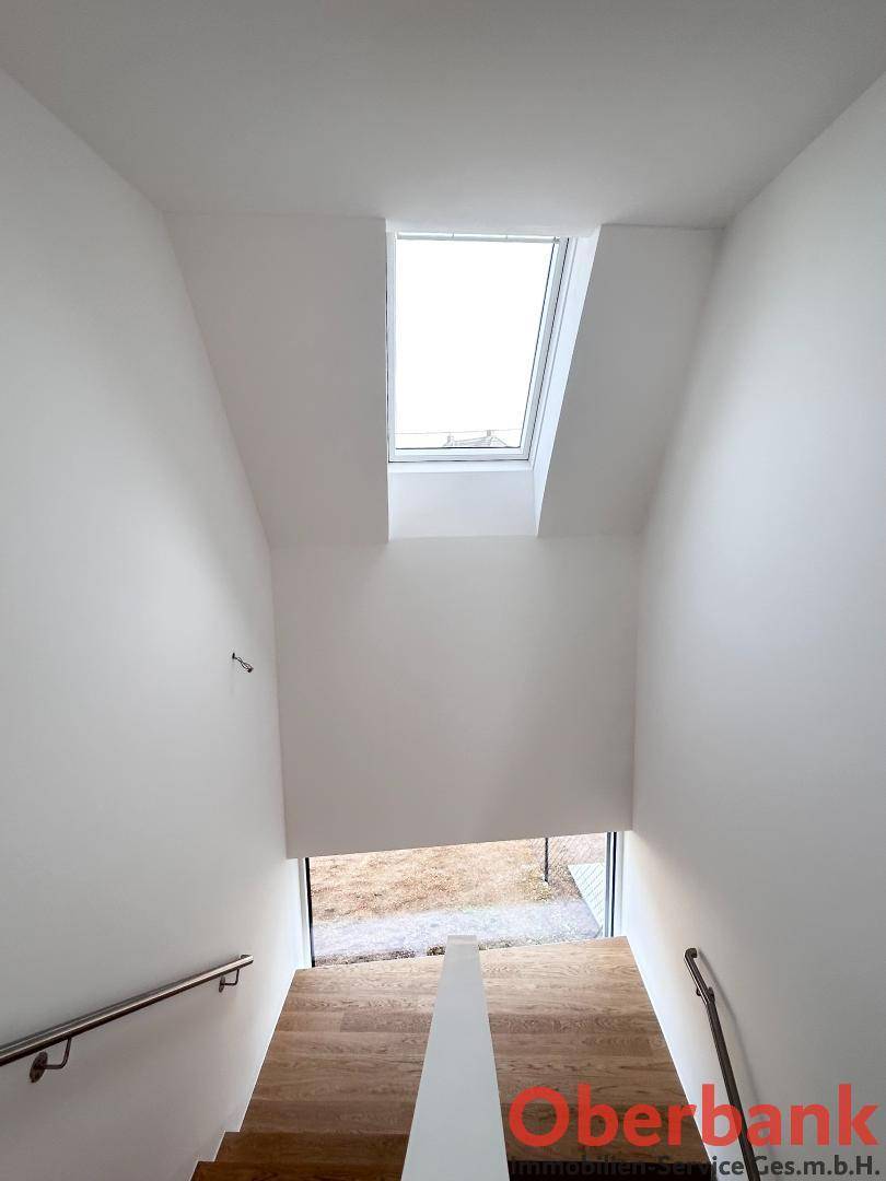 Heller Treppenaufgang mit Holzstufen, Edelstahlgeländer und einem Dachfenster für Tageslicht.
