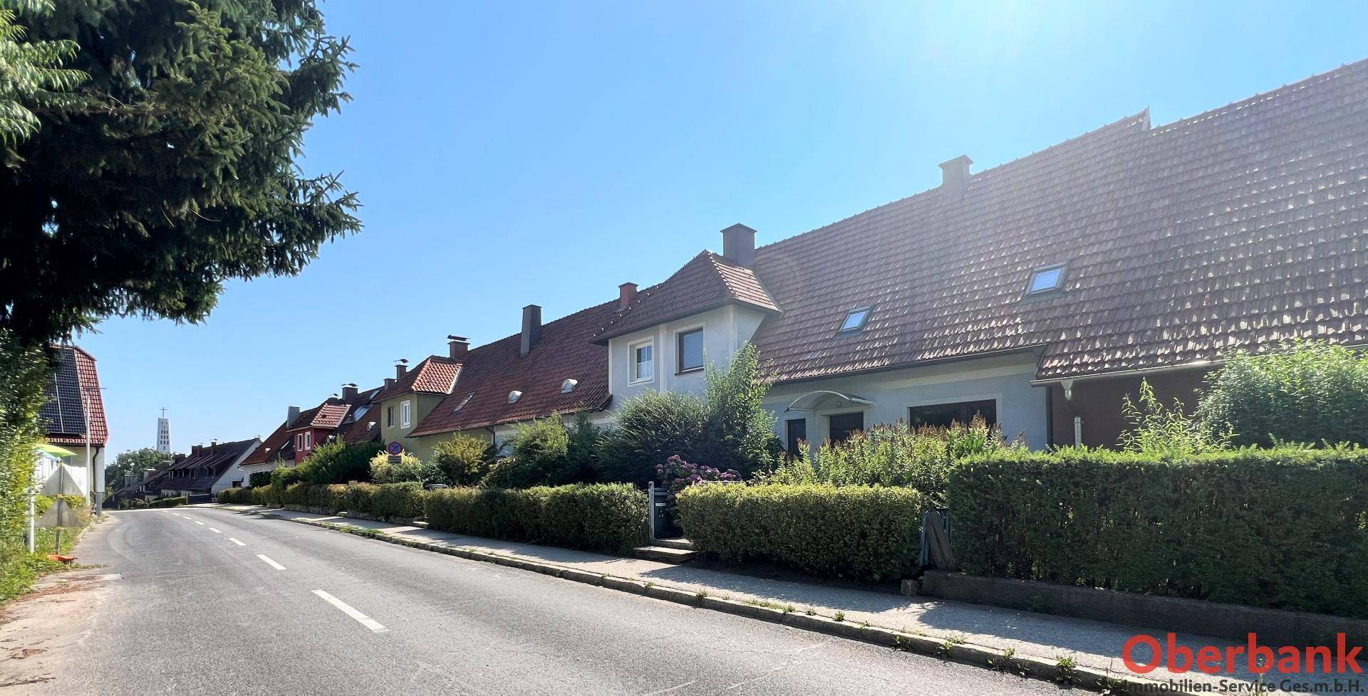 Ruhige Wohnstraße mit gepflegten Häusern und grünen Hecken unter strahlend blauem Himmel.