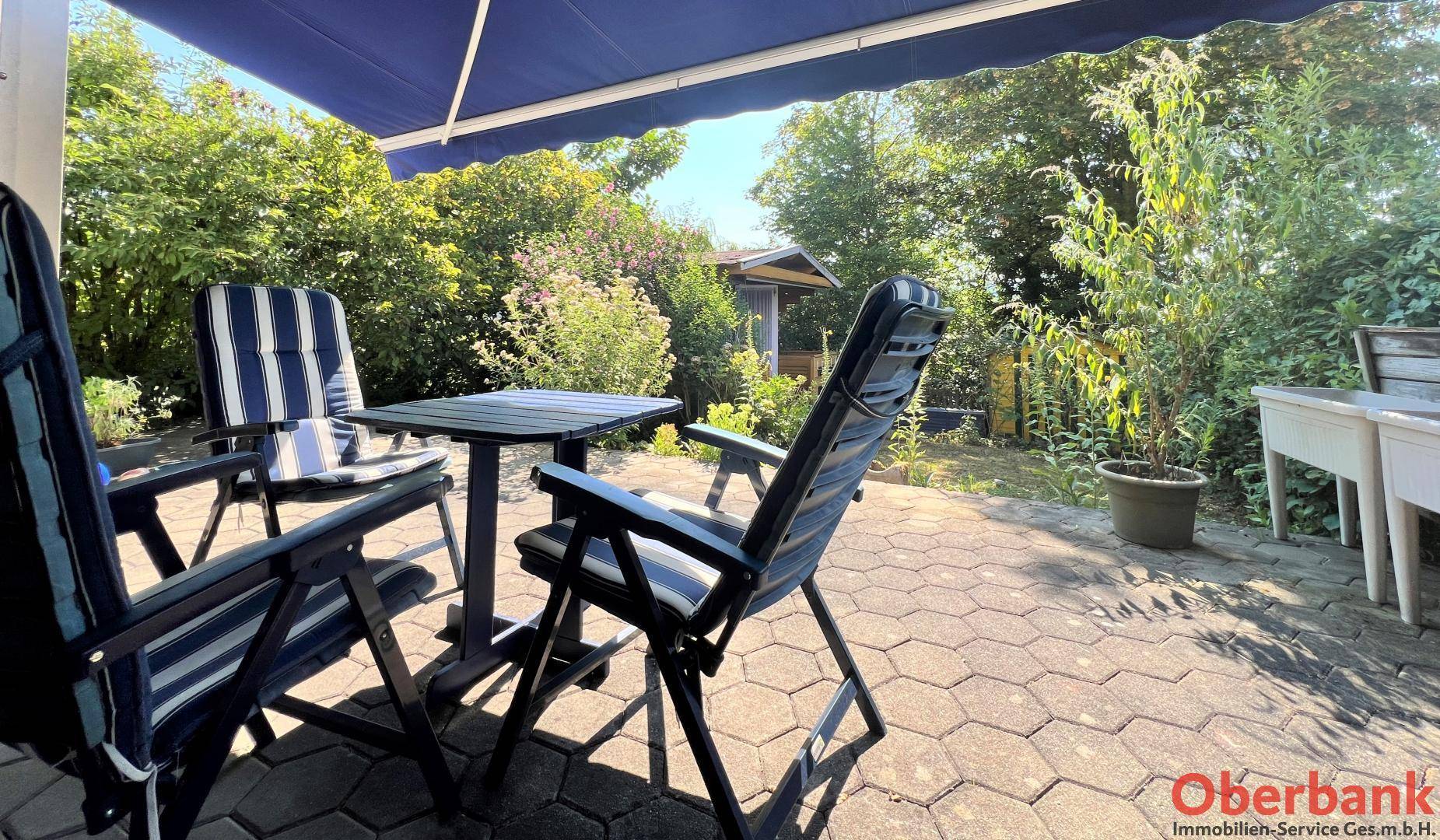 Gemütlicher Terrassenbereich mit blauer Markise und bequemen Gartenstühlen, umgeben von Pflanzen.