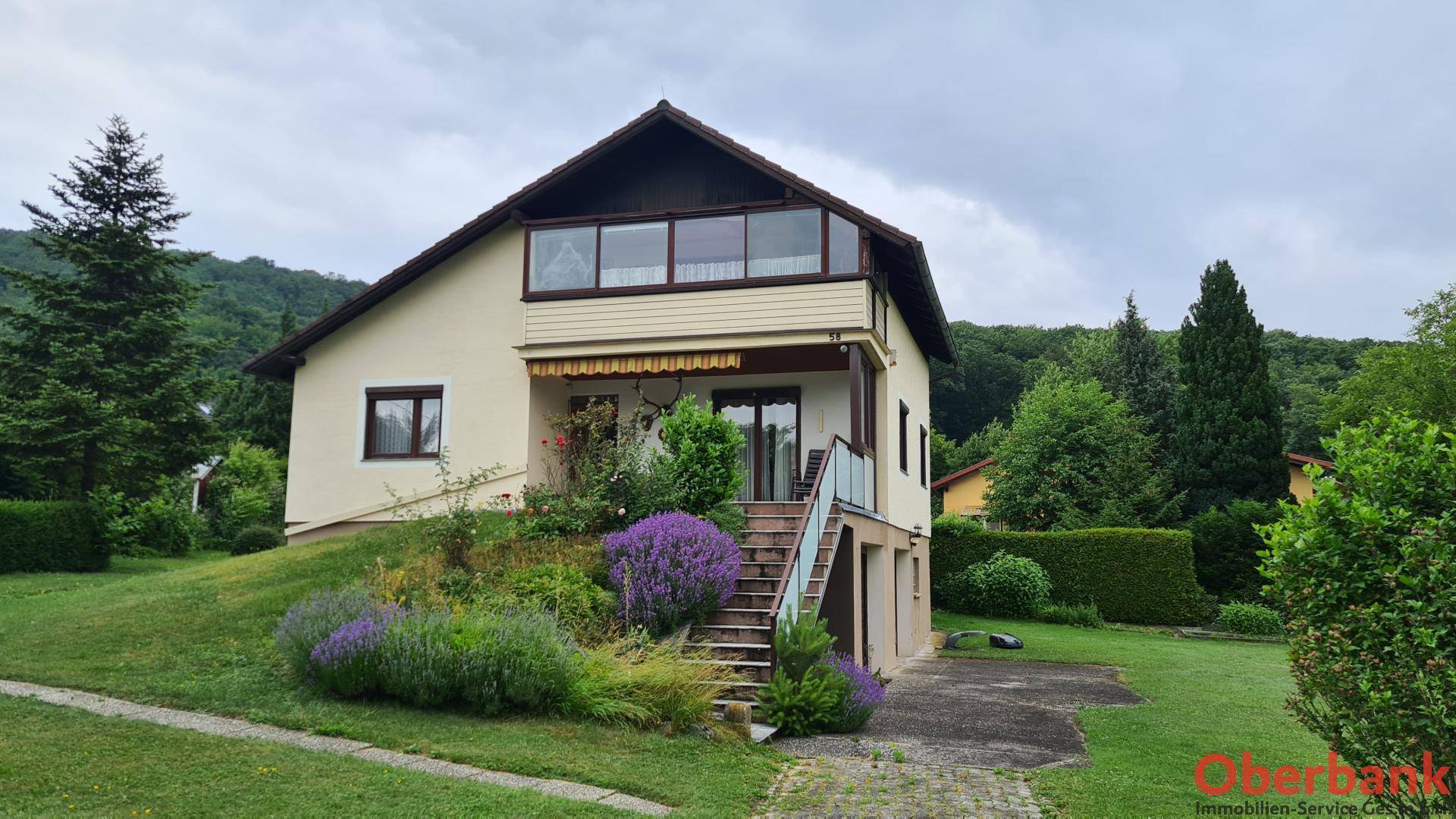 Freistehendes Haus mit Garten, Außentreppe und Wintergarten vor bewaldetem Hintergrund.
