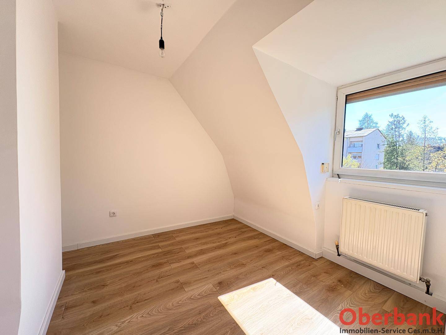 Helles Schlafzimmer mit Holzboden, Dachschrägen und einem Fenster mit Blick ins Grüne.