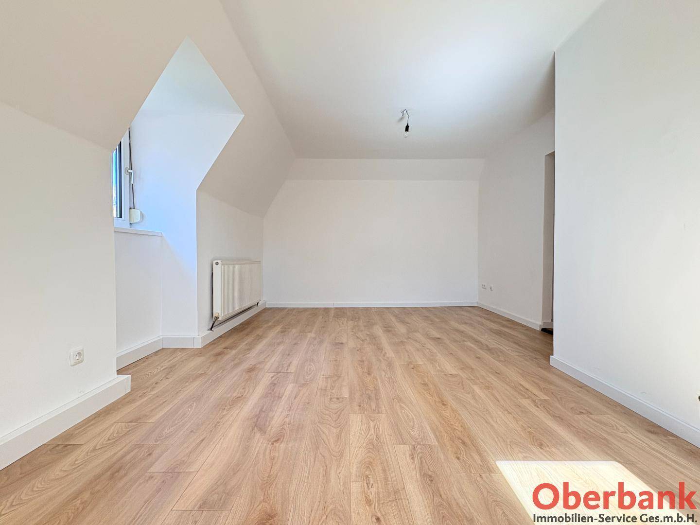 Geräumiges Dachzimmer mit Holzboden, Dachschrägen und einem Heizkörper unter dem Fenster.