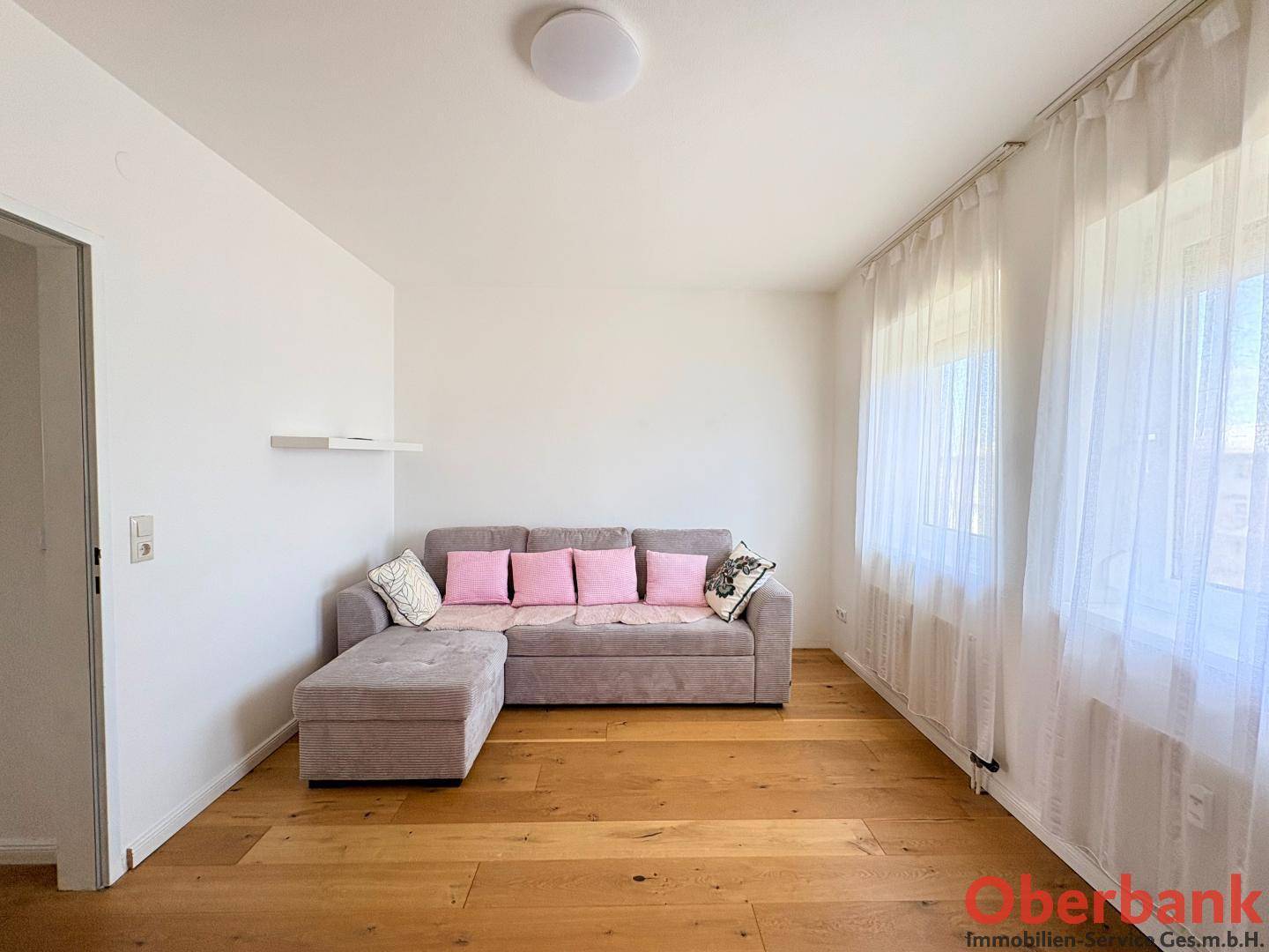 Gemütliches Wohnzimmer mit grauem Sofa, Holzboden und hellen Vorhängen am Fenster.