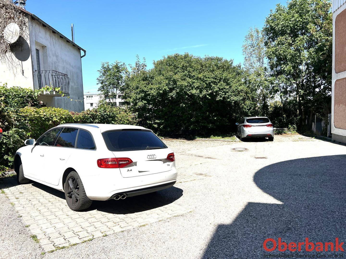 Außenparkplatz mit gepflasterter Fläche und Bäumen im Hintergrund, mit parkenden Autos.