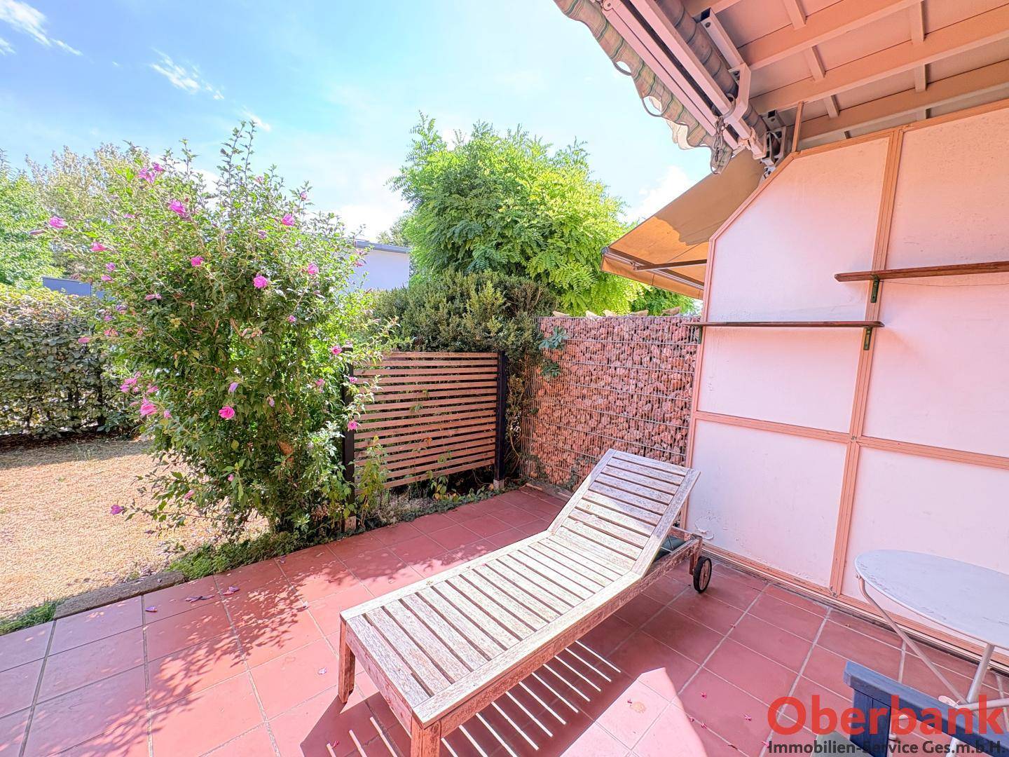Sonnige Terrasse mit Liegestuhl und Blick auf den grünen Gartenbereich mit Sträuchern und Bäumen.