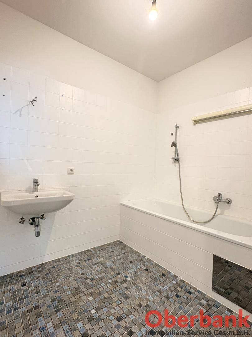 Badezimmer mit weißen Wandfliesen, Mosaikboden, Waschbecken und Badewanne mit Duschfunktion.
