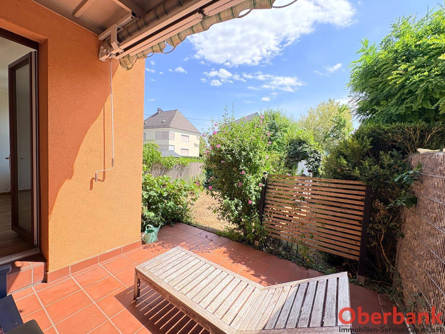 Helle Terrasse mit Liegestuhl und Zugang zum Garten, umgeben von grünen Pflanzen und Hecken.