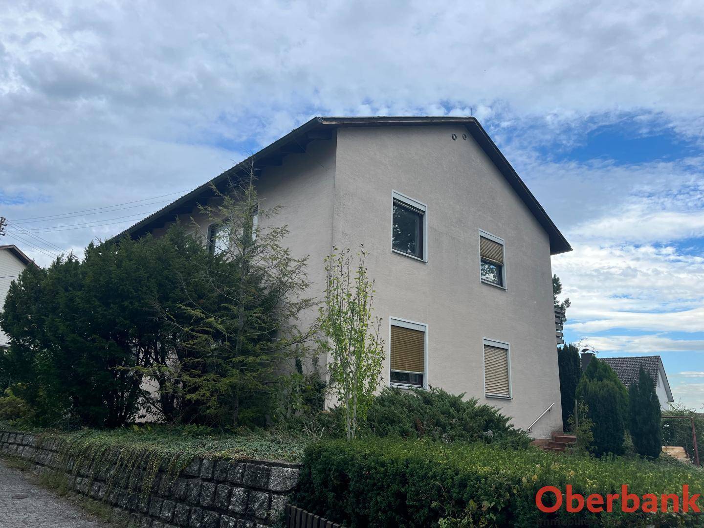 Seitenansicht des Hauses mit Gartenbereich und Rollläden an den Fenstern unter bewölktem Himmel.