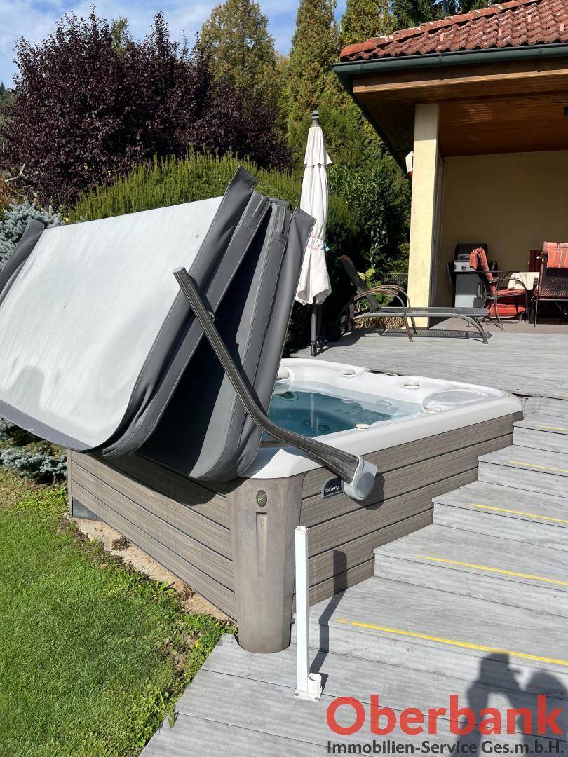 Whirlpool auf der Holzterrasse mit geöffneter Abdeckung, umgeben von grüner Bepflanzung.