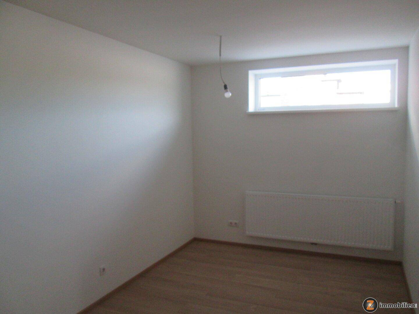 Schlichter, heller Raum mit einem hohen Fenster, Heizkörper und Laminatboden.