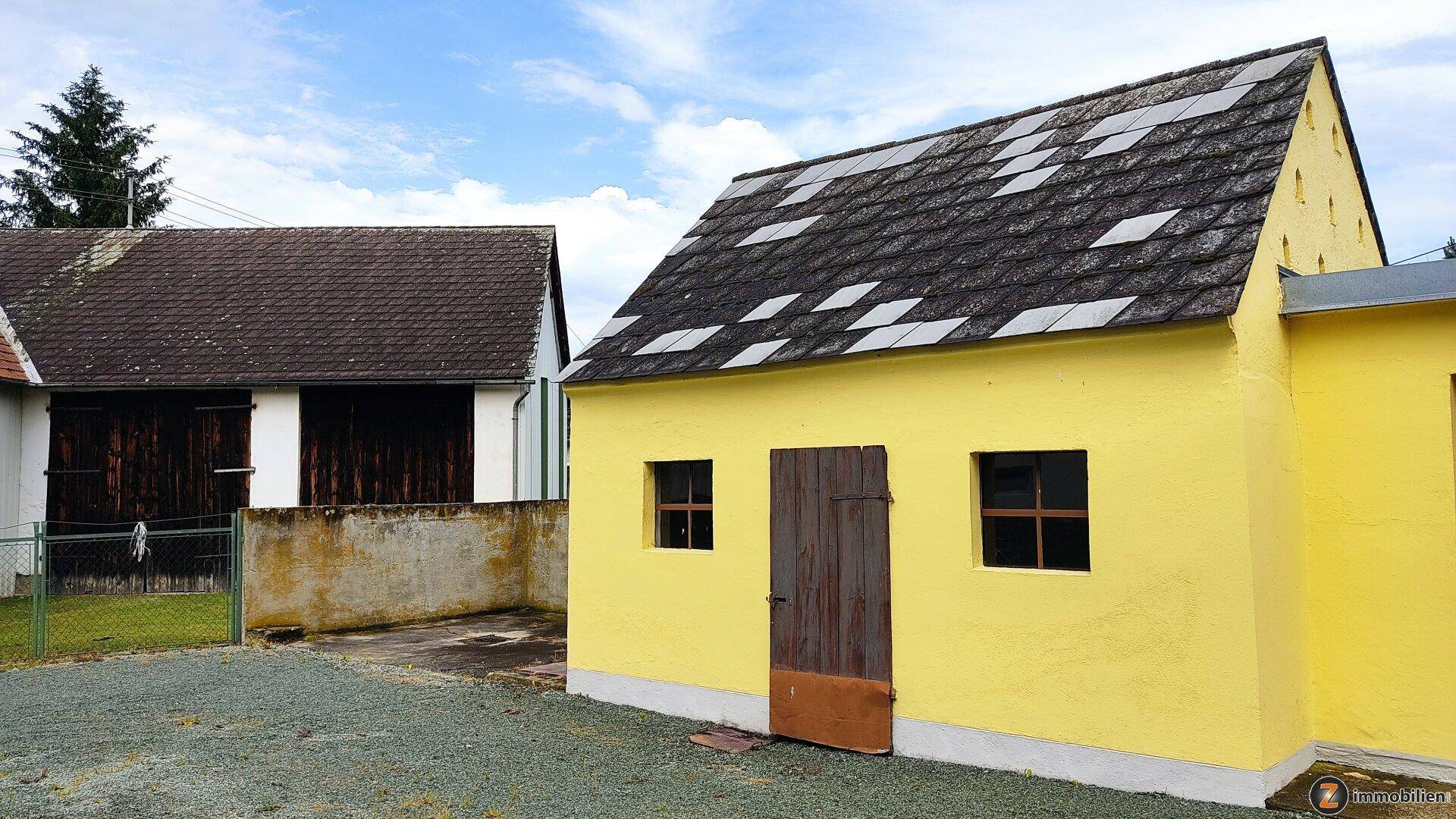 Gelbes Nebengebäude mit Holztür und Fenstern, daneben ein weiteres Gebäude mit dunklem Dach.