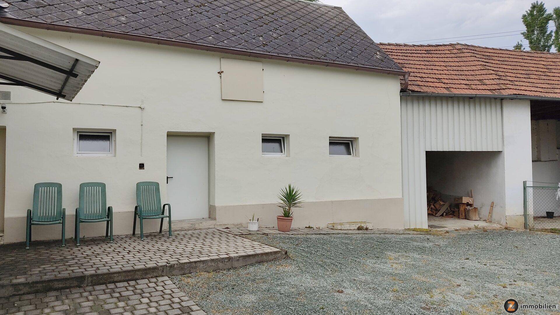 Gepflasterter Bereich vor einem Nebengebäude mit einfachen Gartenstühlen und einem Topfbaum.