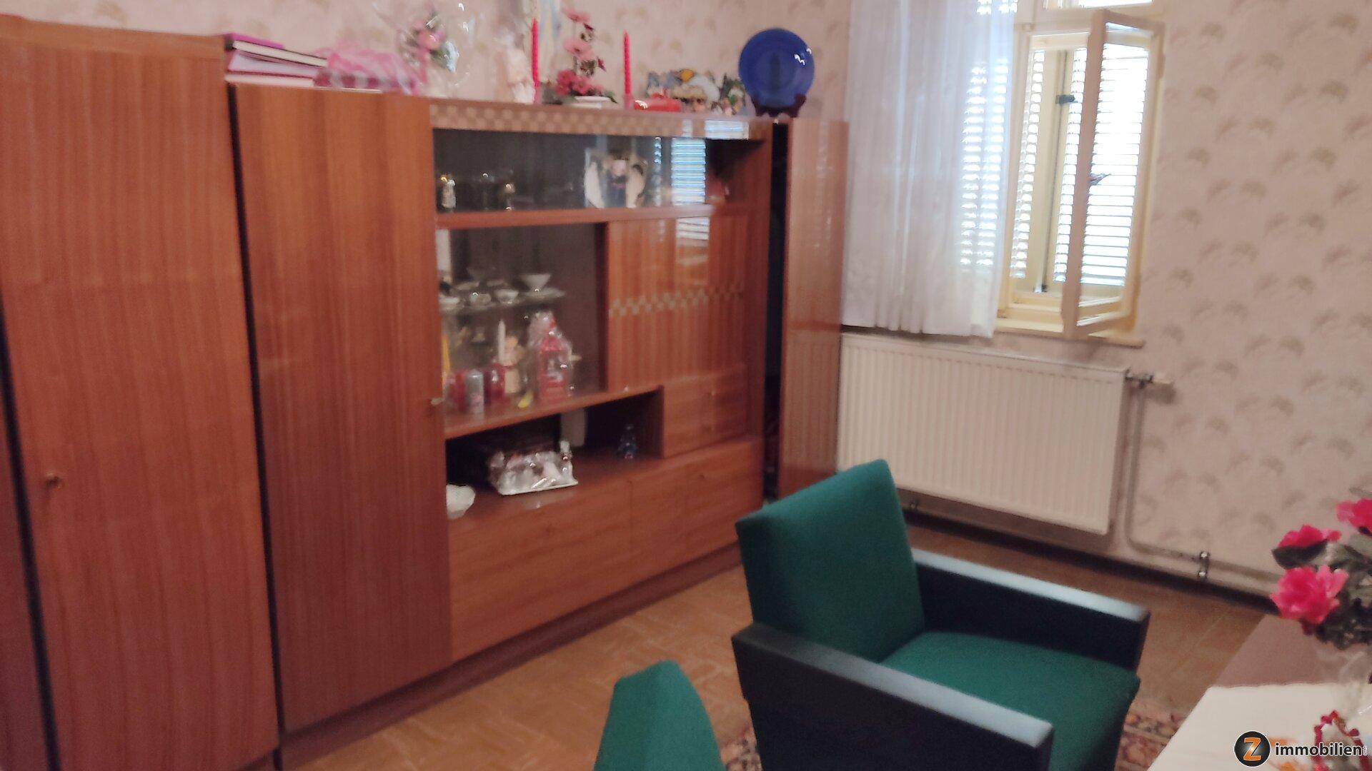 Wohnzimmer mit gemusterter Tapete, Holzmöbeln und einem grünen Sessel.