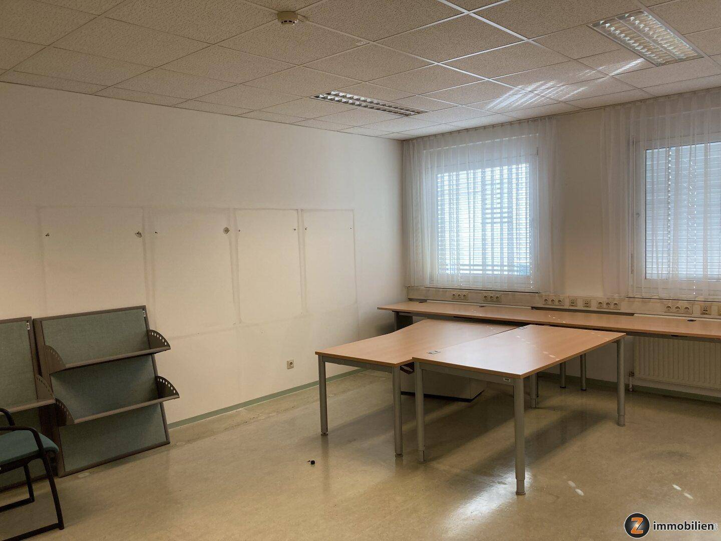 Helles Büro mit mehreren Schreibtischen, großen Fenstern und Vorhängen, ideal für Teamarbeit.
