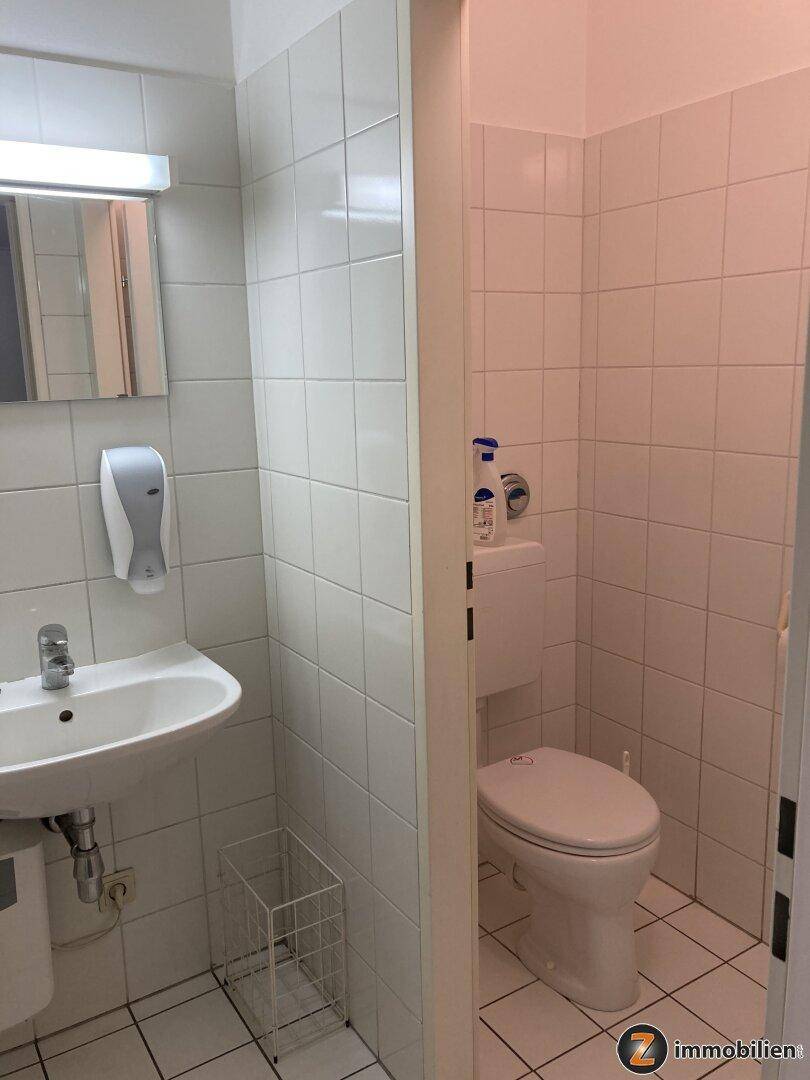 Saubere Toilette mit weißen Fliesen, Waschbecken und Spiegel, funktional und gut gepflegt.