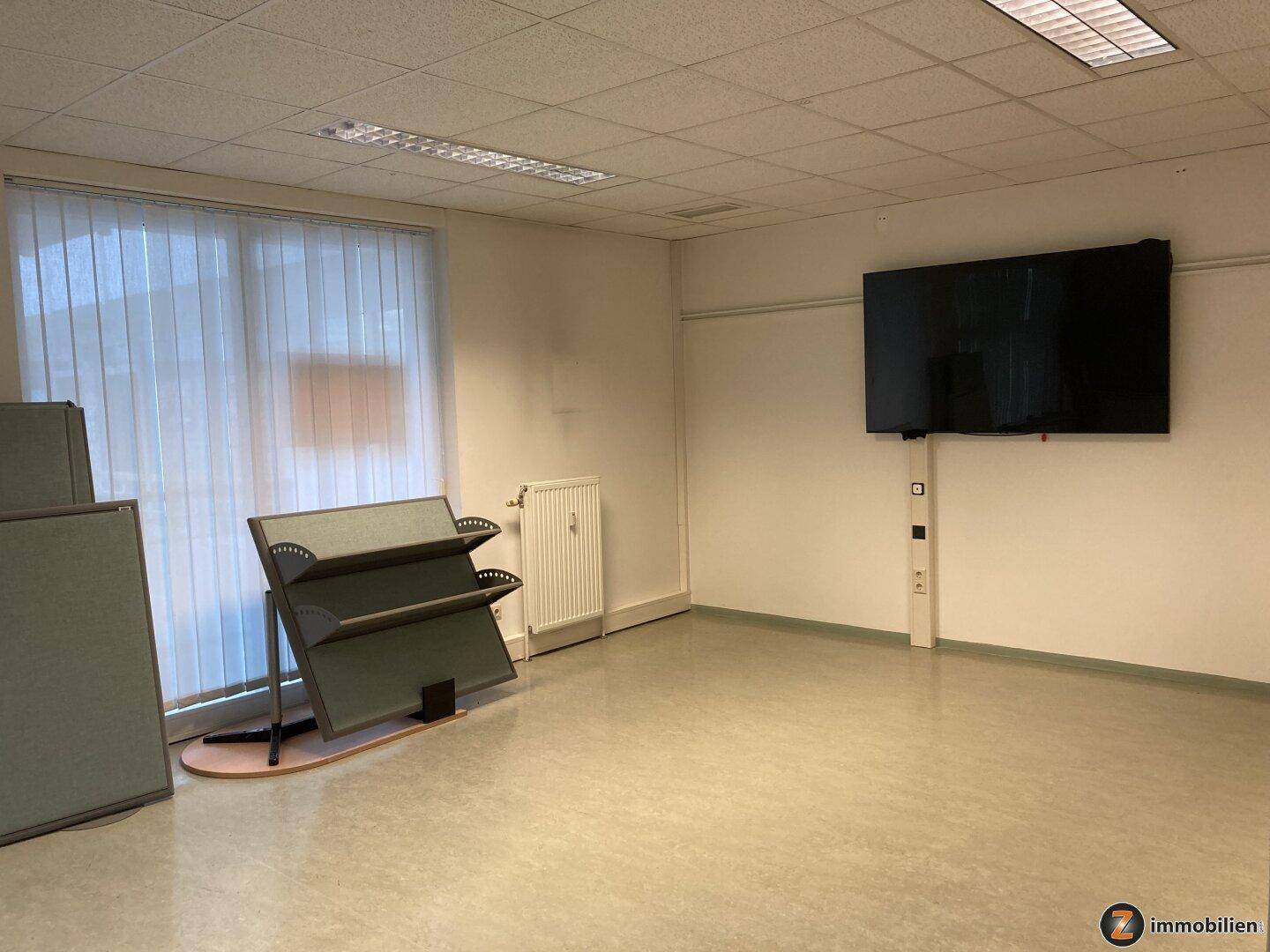 Büro mit großem Fenster, vertikalen Jalousien und einem Flachbildschirm an der Wand, bietet viel Tageslicht.