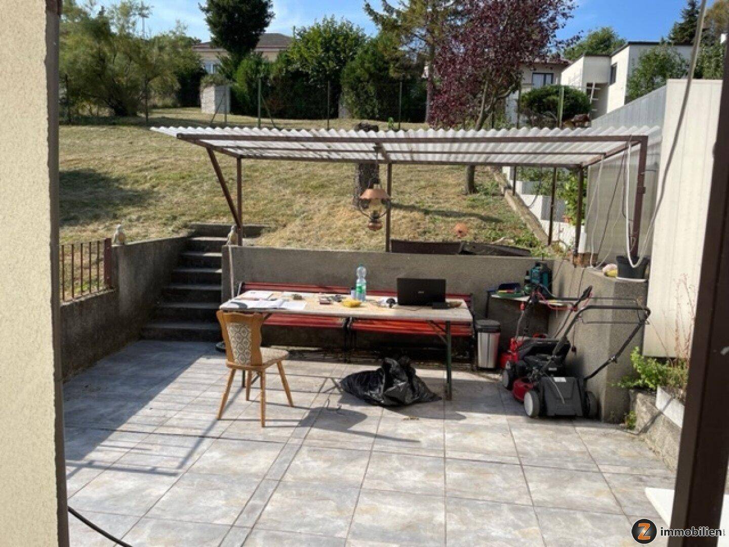 Überdachte Terrasse mit Sitzgelegenheiten, Tisch und Gartengeräten, angrenzend an eine Rasenfläche.