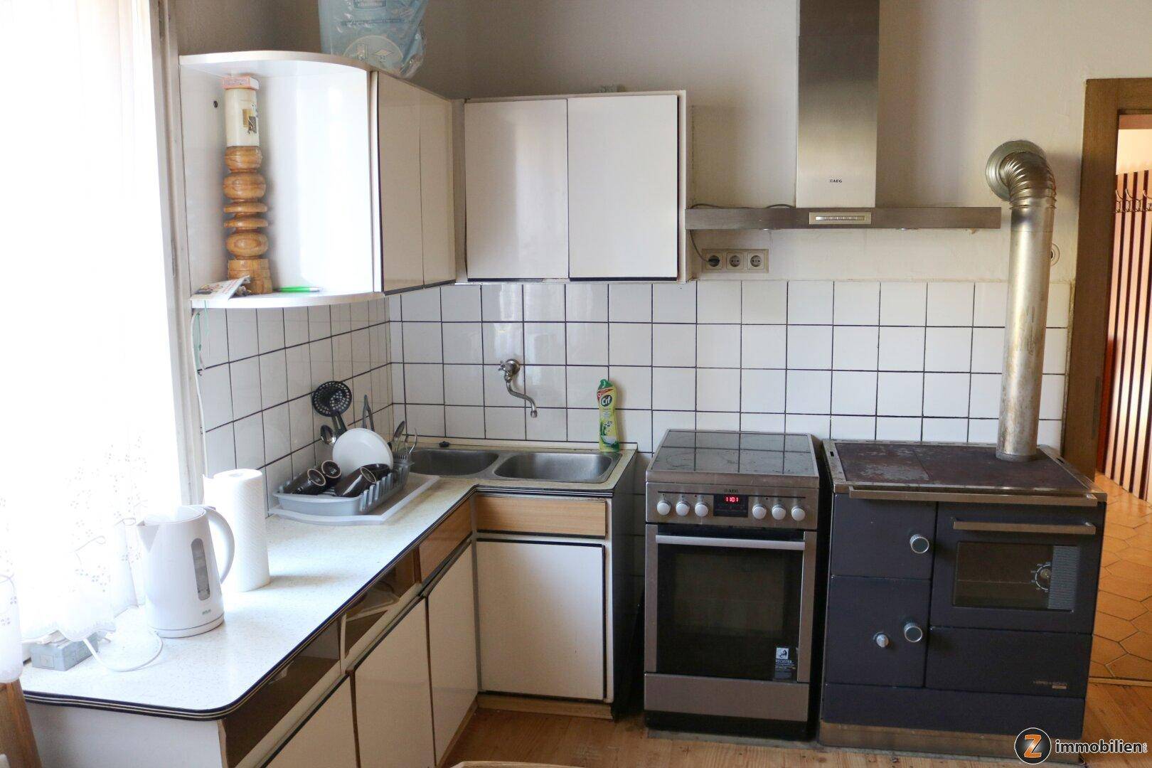 Kleine Küche mit weißen Hängeschränken, Fliesenrückwand und einem traditionellen Holzofen.