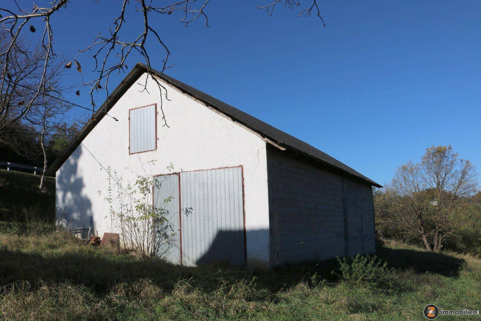 Ein weiß getünchtes Nebengebäude mit Satteldach, Holztoren und einem Fensterladen in ländlicher Umgebung.