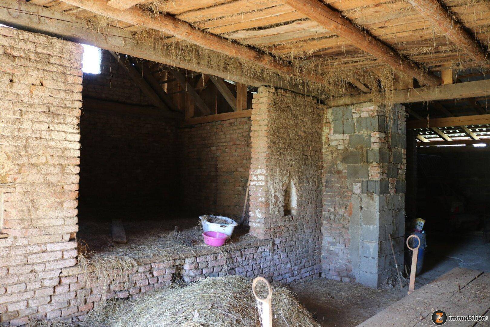 Rustikaler Innenraum eines Nebengebäudes mit sichtbaren Holzbalken, Ziegelwänden und Stroh auf dem Boden.