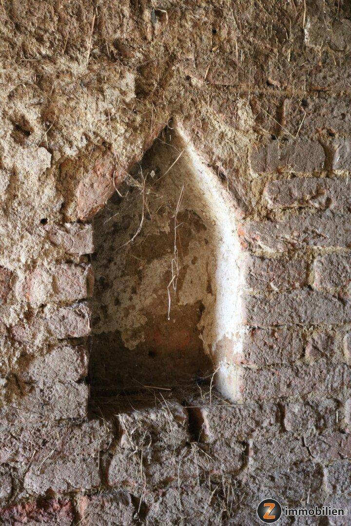 Eine alte, bogenförmige Nische in einer groben Ziegelwand mit Putzresten und Spinnweben.