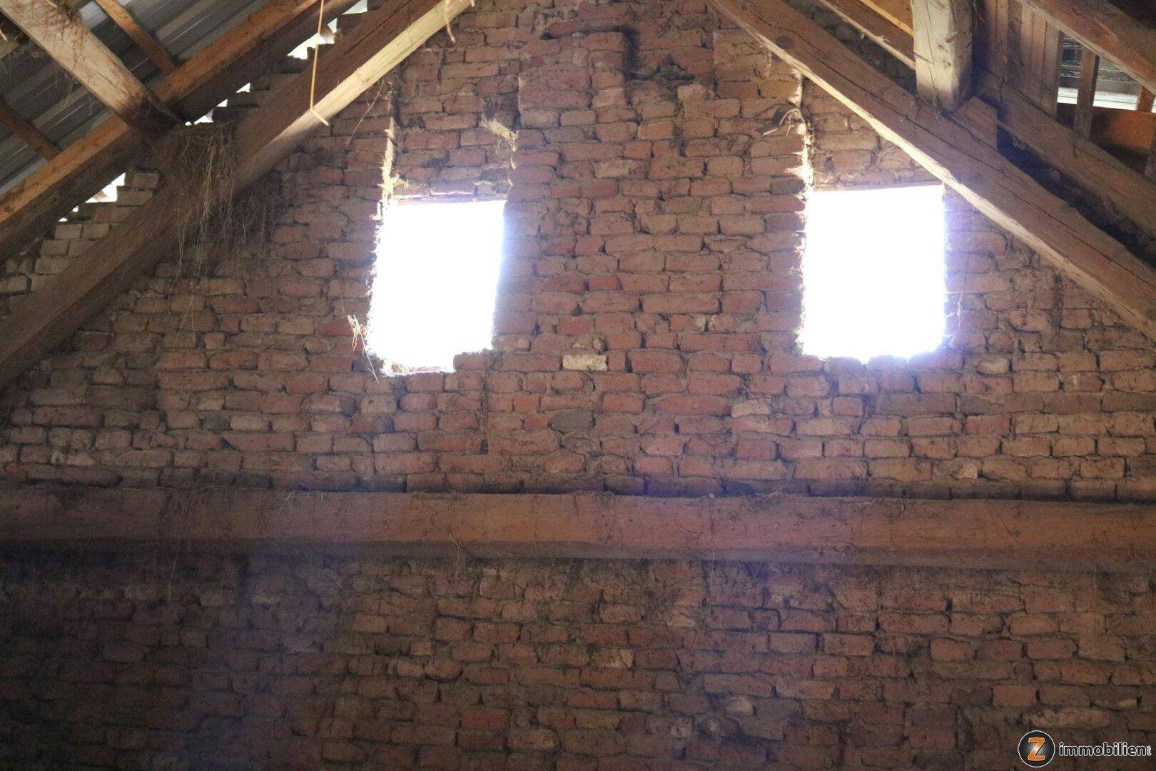Innenansicht eines alten Nebengebäudes mit Ziegelwänden, Holzdachstuhl und zwei kleinen Fenstern.