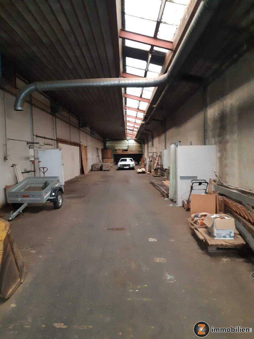 Langer Gang in einer Industriehalle mit Betonboden, Oberlichtern und einem Fahrzeug im Hintergrund.