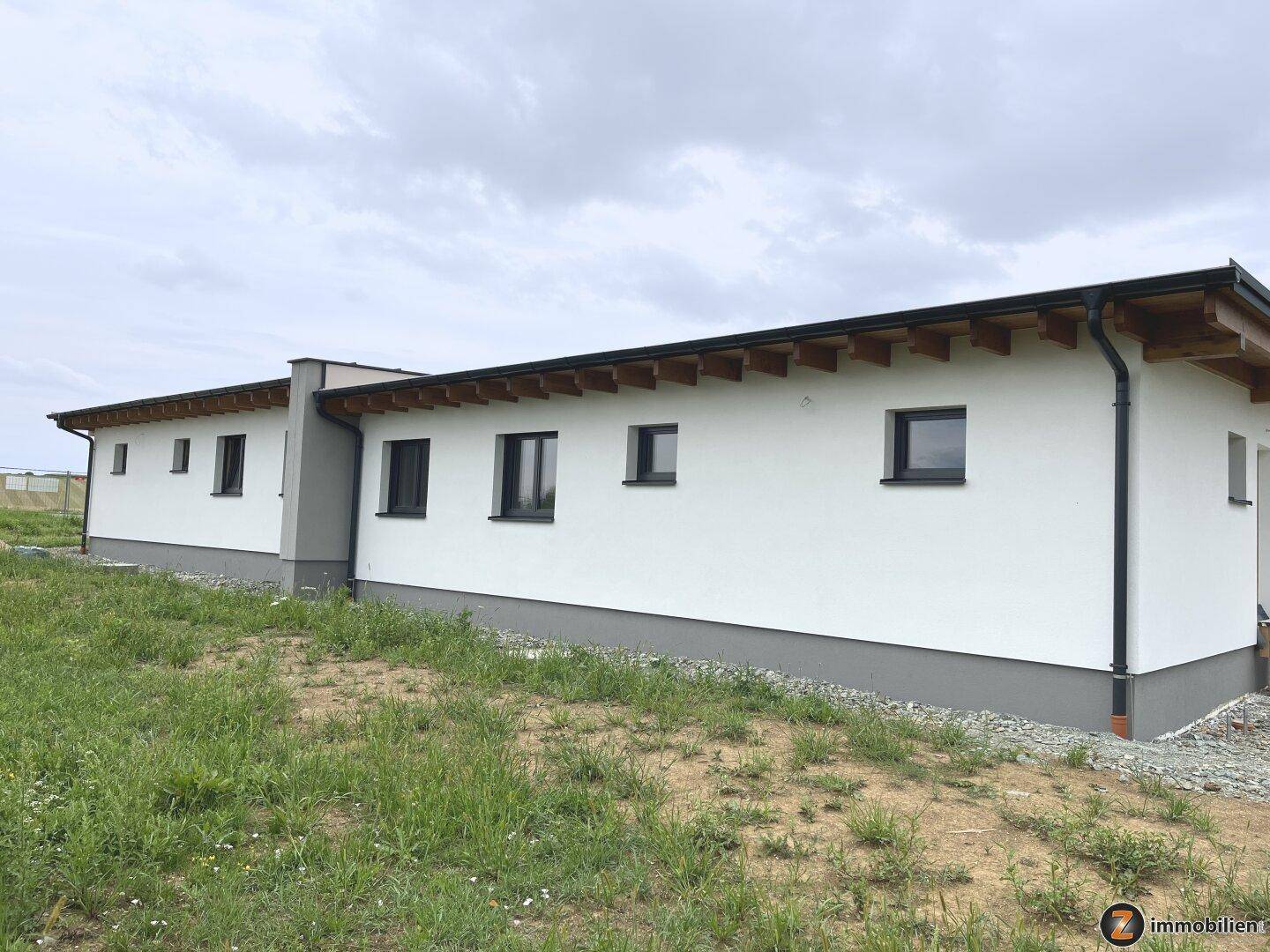 Seitenansicht eines modernen Neubaus mit weißer Fassade und dunklen Fenstern unter bewölktem Himmel.