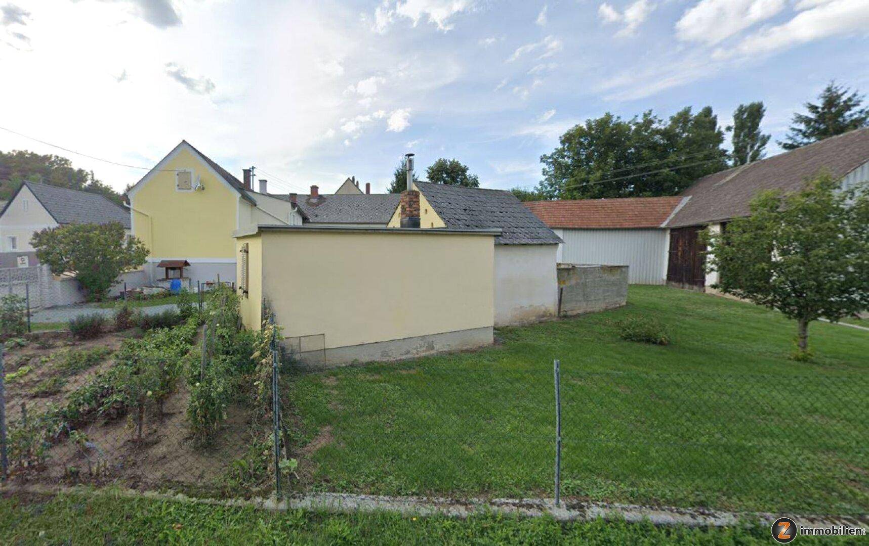 Hinterhof mit Rasenfläche, Gemüsegarten und mehreren Nebengebäuden unter bewölktem Himmel.