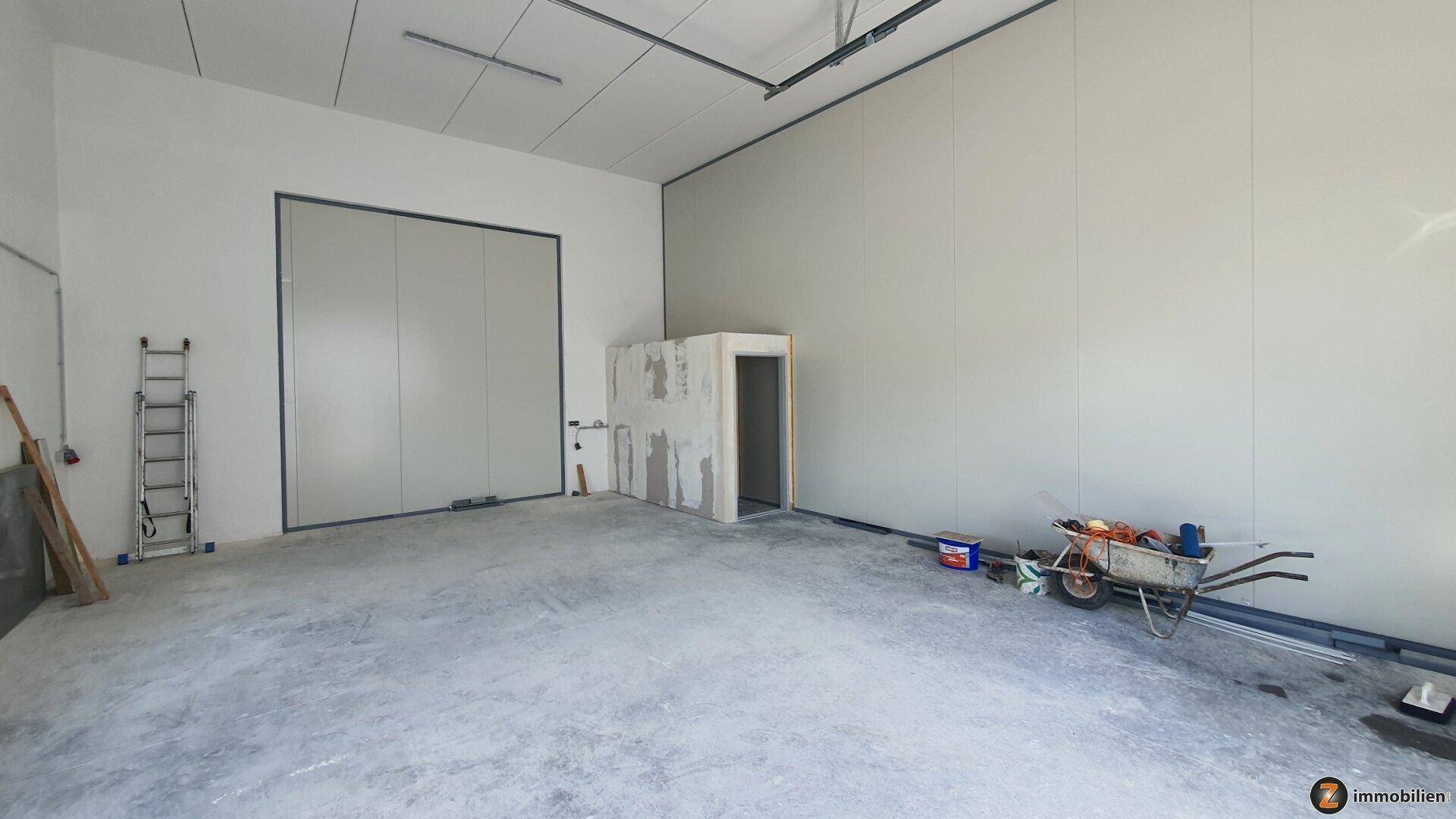 Große, leere Lagerhalle mit Betonboden, hellgrauen Wänden und einer unfertigen Raumeinheit.
