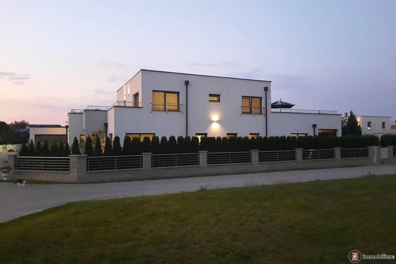 Ansicht der modernen Wohnhausfassade mit großen Fenstern und gepflegter Hecke am Abend.