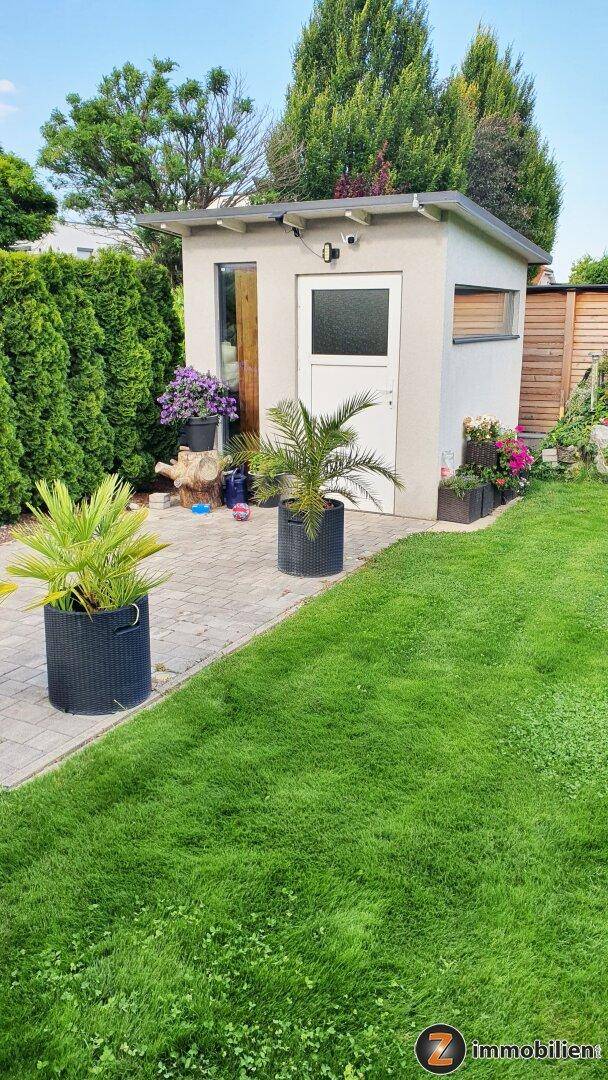 Kleines Gartenhaus mit gepflastertem Weg, umgeben von Rasen und Pflanzen.