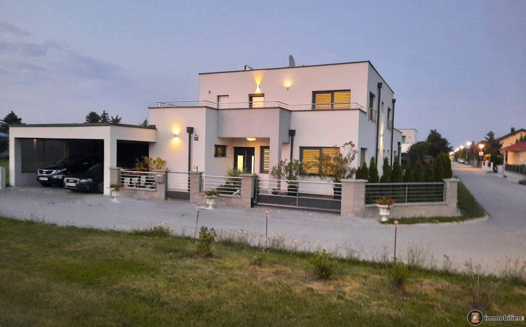 Moderne Wohnhausfassade mit beleuchtetem Eingangsbereich und Carport in der Abenddämmerung.