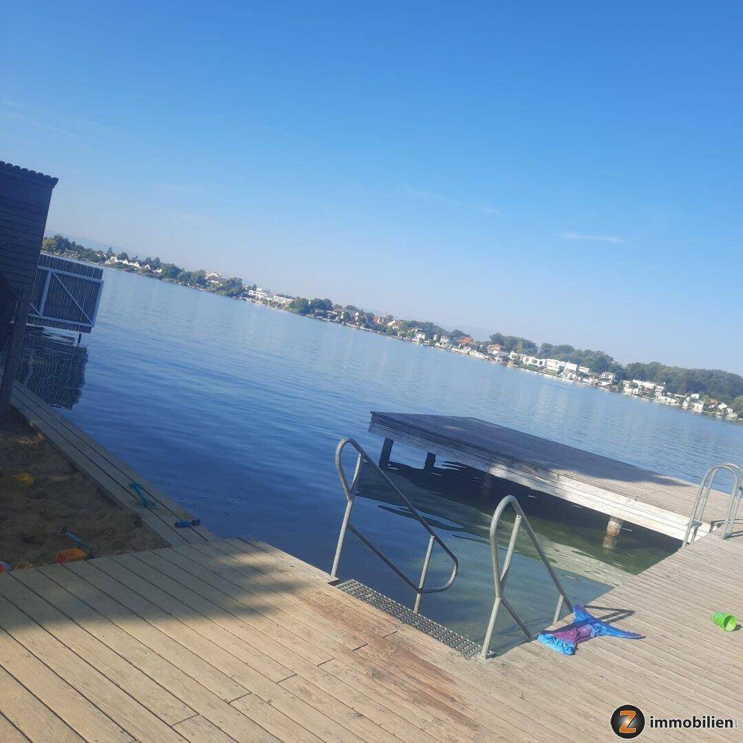 Holzsteg am Seeufer mit klarem Wasser und Häusern am gegenüberliegenden Ufer.