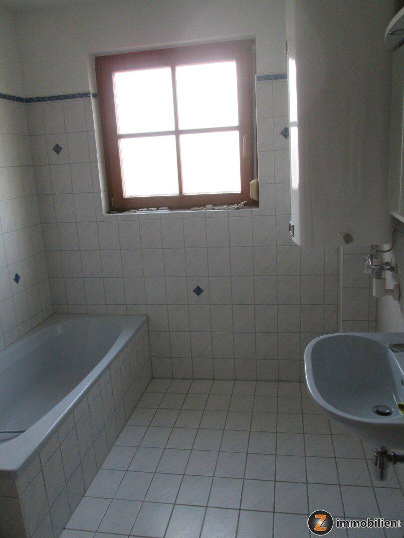 Badezimmer mit Badewanne, Waschbecken und Fenster, gefliest in hellem Grau.