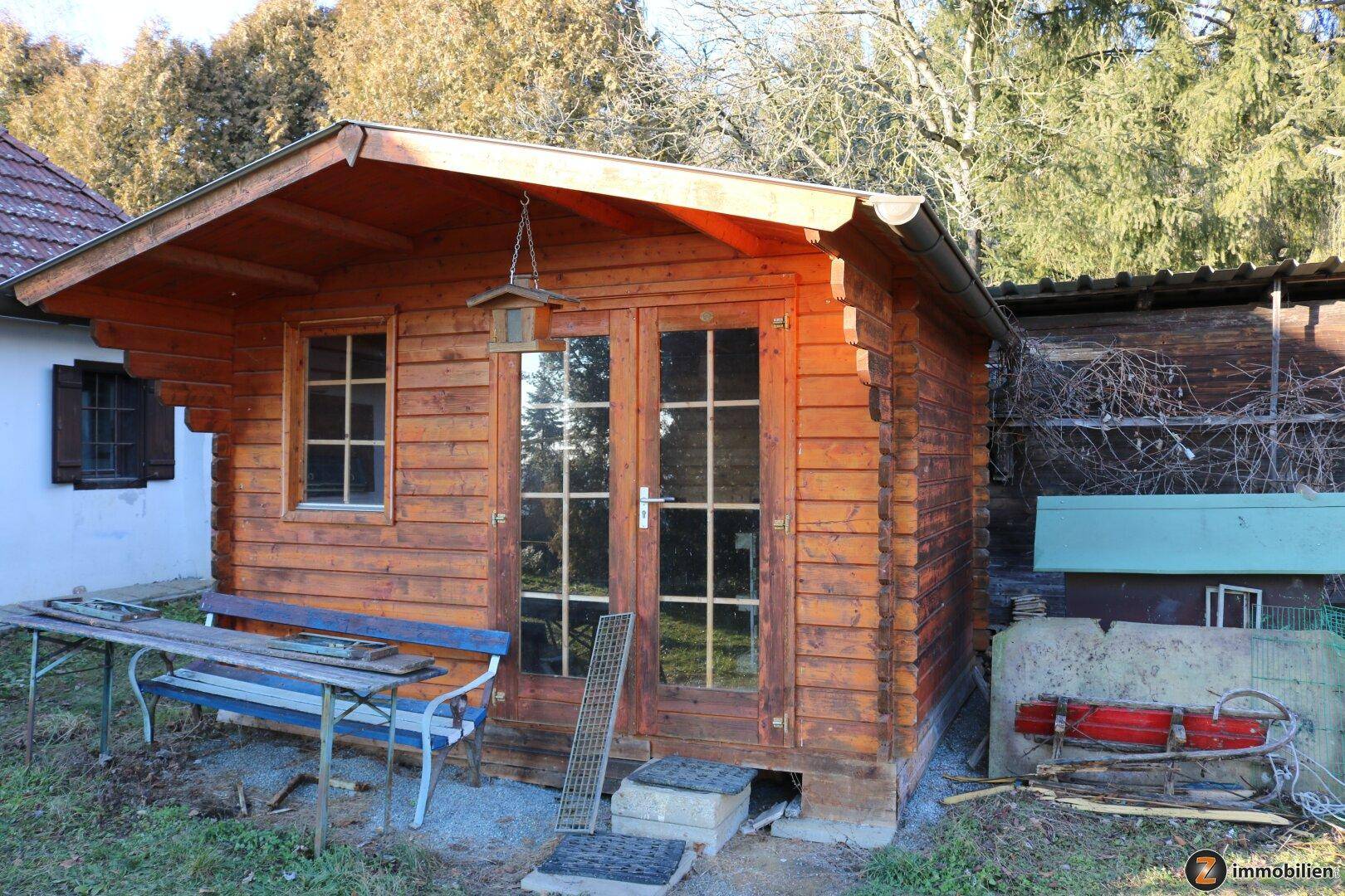 Rustikaler Holzschuppen mit Fenstern und Doppeltür, umgeben von Gartenwerkzeugen.