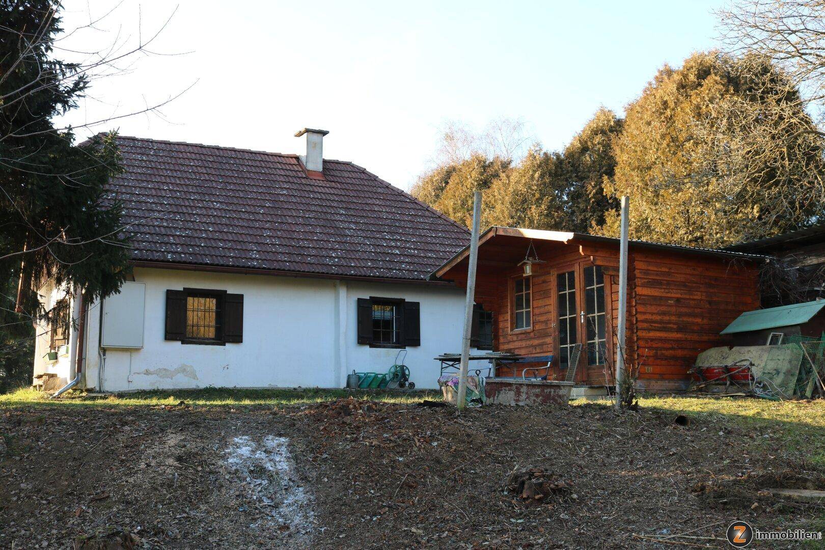 Landhaus mit angrenzendem Holzschuppen und Gartenbereich unter blauem Himmel.