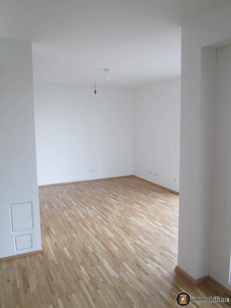 Leerer Raum mit Holzboden, weißen Wänden und offener Tür zu einem weiteren Bereich.