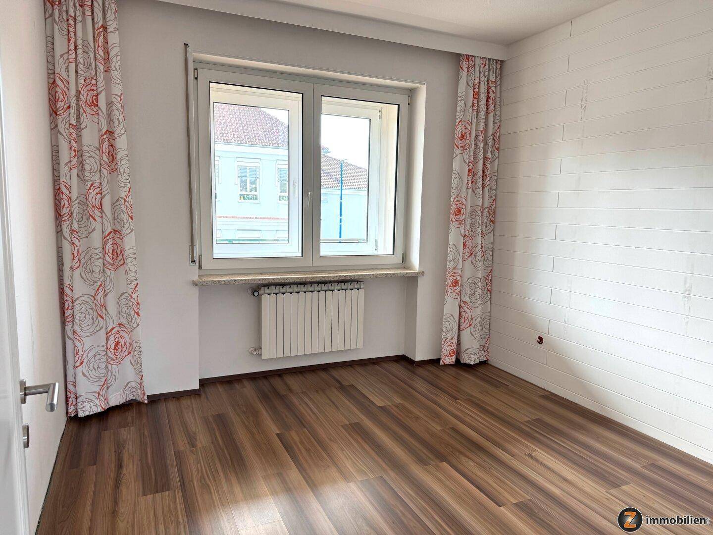 Schlafzimmer mit Laminatboden, Fenster und dekorativen Vorhängen.
